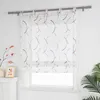 A sheer Roman blind with a curved pattern hangs in front of a window. Decorative elements like a vase with branches and a framed picture are visible on a mantelpiece to the left, and a potted plant is on the right. Produktbild-Vorschau