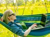 A woman is relaxing in a hammock outdoors, using a laptop. Produktbild-Vorschau