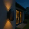 An outdoor wall lamp is mounted on a textured wall, casting light upwards and downwards. The background shows a house with large windows and a garden at dusk. Produktbild-Vorschau