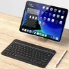A black portable Bluetooth keyboard and a white stylus are placed on a wooden desk next to a tablet displaying a home screen with various app icons. Náhled obrázku produktu