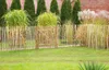 A wooden picket fence with a gate in a garden setting, with grass in the foreground and trees and a building in the background. Produktbild-Vorschau
