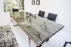 A dining table with glass top and wooden base, set in a modern dining room with chairs and a rug. Produktbild-Vorschau