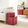 Two red storage boxes are stacked on a rug next to a cream-colored sofa. A wooden shelf unit with plants and more storage boxes is in the background. Produktbild-Vorschau