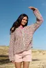 A woman stands in a sandy, outdoor environment under a clear blue sky, wearing a colorful knitted cardigan and pink shorts. Produktbild-Vorschau