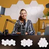 A woman is sitting at a desk with a microphone in front of her, with hexagonal acoustic panels on the wall behind her. Below the main image are three arrangements of the hexagonal panels. Produktbild-Vorschau