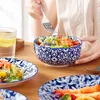 A person is eating a salad from a blue and white patterned bowl, with other plates and a glass of water on a wooden table. Produktbild-Vorschau