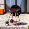A black charcoal grill with silver legs and wheels, placed on a paved patio in front of a large window. Náhľad obrázku produktu