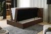 A brown boxspring bed with its storage compartment open, revealing the mattress inside. The bed is in a room with a closet in the background and a lamp on the right. Produktbild-Vorschau