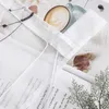 A close-up view of a white fabric with a gray embroidered pattern, layered over a book and a knitted blanket, with a cup of coffee and a dried leaf in the background. Produktbild-Vorschau