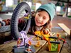 A young girl is playing with a LEGO stunt park set, which includes a motorcycle, a spider cage, and a minifigure. The set is arranged on a wooden table, with a large tire serving as a ramp. Podgląd zdjęcia produktu