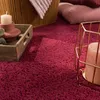 A close-up view of a plush, red rug with decorative items on it, including a candle in a holder and a wooden tray. Produktbild-Vorschau