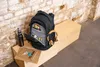 A black backpack is open and filled with various items, including headphones, a water bottle, and a notebook, placed on a wooden bench against a brick wall. Produktbild-Vorschau
