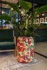 A tall, decorative plant pot with a floral pattern sits on a rug next to a green armchair in a modern interior setting. Produktbild-Vorschau