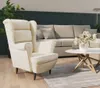 A cream-colored wingback armchair is placed in a living room setting with a sectional sofa and a coffee table with flowers. Anteprima dell’immagine prodotto