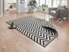 A chevron patterned rug is laid out on a wooden floor in a living room setting. A grey armchair with a blanket is visible in the background. Voir les images du produit
