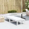 An outdoor patio scene with a white wooden sun lounger, a small wooden side table, planters with grass, and a large umbrella pole. Náhled obrázku produktu