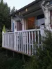 A view of a blue house with a white railing on a deck, surrounded by greenery. Produktbild-Vorschau