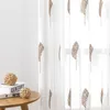 A close-up view of a sheer curtain with a leaf embroidery pattern, placed next to a small wooden table with decorative balls and books. Produktbild-Vorschau