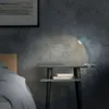 A teal desk lamp with a clip base is attached to a small gray shelf, illuminating two books. The scene is set next to a bed with white bedding, against a textured dark gray wall. Produktbild-Vorschau