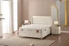 A bedroom scene featuring a large bed with a quilted headboard and mattress, a bedside table with a lamp, and sheer curtains. Produktbild-Vorschau
