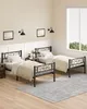 Two twin beds with black metal frames and white bedding are set up in a room with cream walls and light wood flooring. There is a window with blinds on the right side of the image. Two framed pictures hang on the wall above the beds, and two wall-mounted l Náhled obrázku produktu