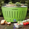 A green LotusGrill charcoal grill with food on it, placed on a wooden table with bread and tomatoes. Náhled obrázku produktu
