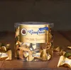 A clear, well-lit studio shot of a round plastic container filled with individually wrapped chocolates, placed on a wooden surface. Náhľad obrázku produktu