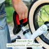 A person's hand is holding the seat of a child's balance bike, with the bike's wheel and frame visible. The background is blurred greenery and pavement. Produktbild-Vorschau