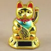 A golden lucky cat figurine with a red collar and teal bib, holding a gold ingot, sits on a round base with a solar panel. Produktbild-Vorschau