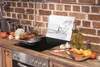 A kitchen counter with various cooking ingredients and utensils, including a cutting board with chopped vegetables, bottles of oil, and a frying pan on a stovetop. A decorative glass panel with a diamond image is placed behind the stovetop. Produktbild-Vorschau