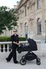 A woman is pushing a black stroller in front of a large, ornate building. Produktbild-Vorschau