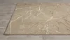 A close-up, angled view of a beige and gray rug with a branch-like pattern, lying on a wooden floor. Produktbild-Vorschau