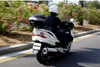 A person riding a white scooter on a road with green bushes and pink flowers. Anteprima dell’immagine prodotto