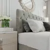 A close-up view of a bed with a tufted headboard, pillows, and a bedside table with flowers and books. Produktbild-Vorschau