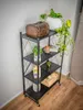 A black metal shelving unit with items on each shelf, placed against a light gray wall and on a wooden floor. Produktbild-Vorschau