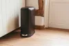 A black pedal bin is placed on a wooden floor next to a white wall and a wooden beam. Produktbild-Vorschau