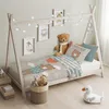 A child's bed with a teepee frame, decorated with fairy lights and framed pictures. The bed is made with bedding featuring bear illustrations. There are stuffed animals and storage baskets nearby. Náhled obrázku produktu