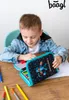 A young boy is sitting at a wooden table, looking down at an open pencil case filled with colored pencils. He is wearing a denim jacket and a white t-shirt. There are also loose colored pencils on the table. Anteprima dell’immagine prodotto