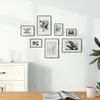 A collection of framed pictures displayed on a white wall above a white cabinet, with a plant and a book on a wooden table in the foreground. Produktbild-Vorschau
