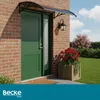 A green front door with a gray awning above it, set against a brick wall. There is a lantern and a planter with red flowers next to the door. The scene is outdoors with a lawn and trees in the background. Náhled obrázku produktu