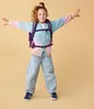 A young girl wearing a colorful cardigan and jeans, with a backpack on, stands with her arms outstretched. Náhled obrázku produktu