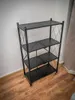 A black metal shelving unit with four perforated shelves is shown against a white wall and on a wooden floor. Produktbild-Vorschau