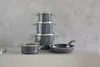 A set of dark gray pots and pans stacked and arranged on a light gray surface against a textured gray background. Anteprima dell’immagine prodotto