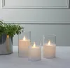 Three LED candles in glass holders are arranged on a table with a plant in the background. Podgląd zdjęcia produktu