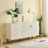 A white sideboard with gold accents and drawers, decorated with plants and a lamp, in a room with a light yellow wall and a rug. Produktbild-Vorschau