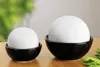 Two white spheres are placed in black bowls on a wooden surface, with a blurred green background. Produktbild-Vorschau