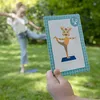 A child is practicing yoga outdoors, mimicking a pose shown on a card held by a hand. Produktbild-Vorschau