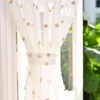 A close-up of a sheer white curtain with small beige pom-pom accents, hanging in front of a window. Produktbild-Vorschau