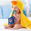 A baby wearing a duck-themed hooded towel is sitting on a bed and playing with a soft fabric book. Anteprima dell’immagine prodotto