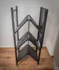 A black metal folding shelf unit with four shelves is shown standing on a wooden floor against a white wall. Produktbild-Vorschau